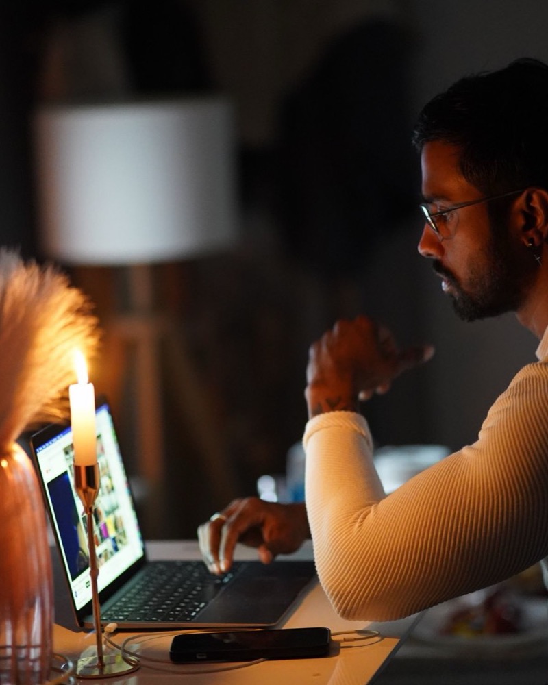 Hombre de perfil absorto frente a la pantalla de un portátil abierto, iluminado por la luz cálida de una vela y una lámpara en su escritorio nocturno, tratamiento de las adicciones comportamentales en adultos en Komit Psicología