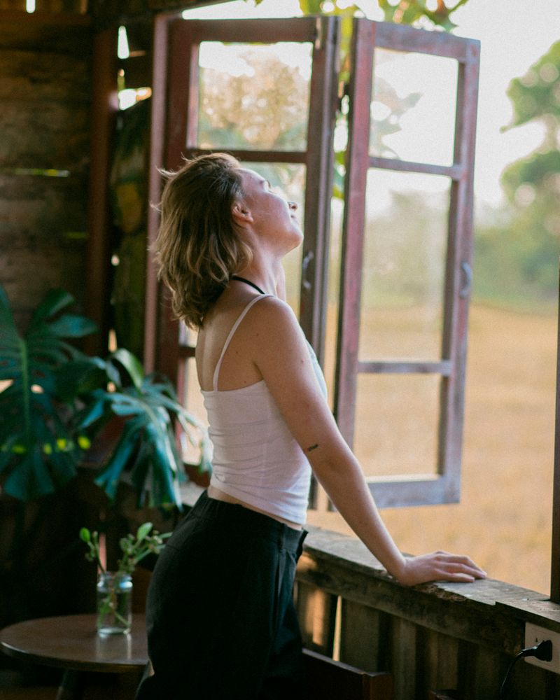 Mujer adulta junto a una ventana abierta con los ojos cerrados y el rostro hacia la cálida luz del atardecer, respirando en calma entre plantas, terapia de la ansiedad en Komit Psicología