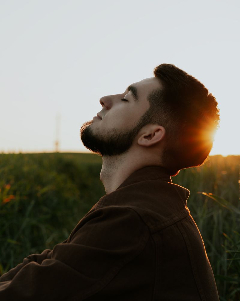 Hombre adulto de perfil con los ojos cerrados respirando en calma bajo la cálida luz dorada del atardecer en un campo verde, terapia del trastorno de conducta alimentaria en adultos en Komit Psicología