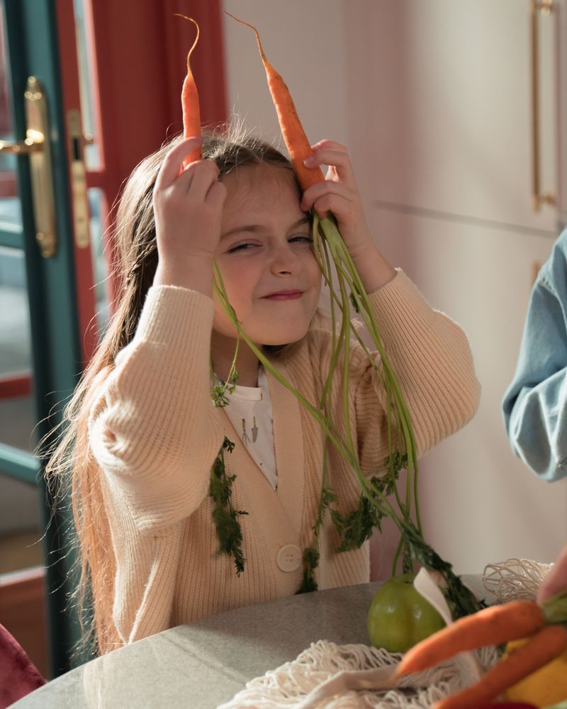 Niña sonriente jugando con zanahorias frescas junto a la mesa de casa con luz natural cálida, terapia de problemas de alimentación en la infancia en Komit Psicología